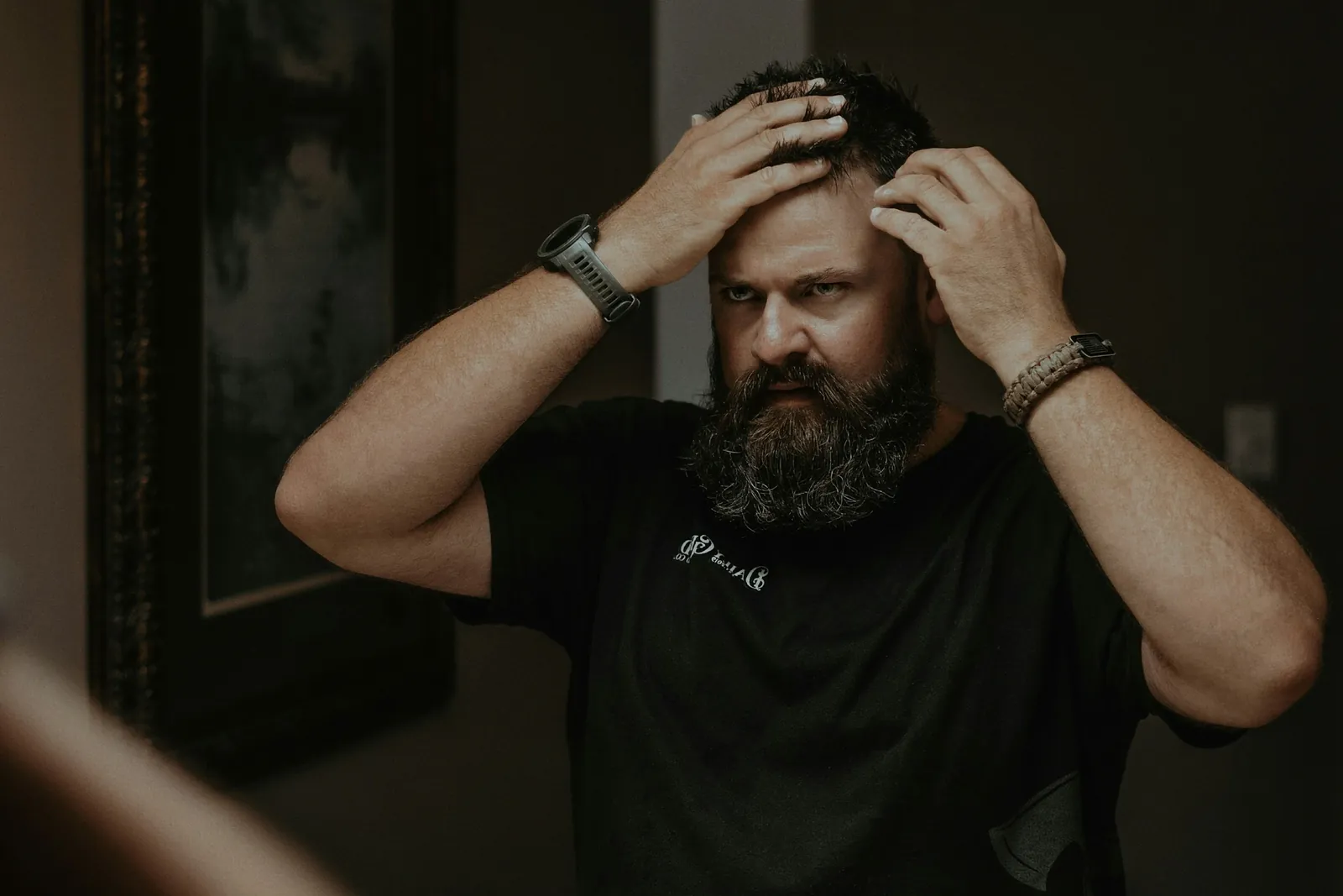 A bearded man in a dim bathroom mirror, both hands pushing back his hair to inspect his hairline.